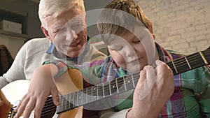 Grandfather is teaching his grandson how to play the guitar. An elderly man is teaching a young fat child to play guitar