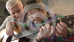 Grandfather is teaching his grandson how to play the guitar. An elderly man is teaching a young fat child to play guitar