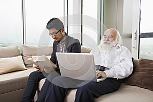 Grandfather using laptop while grandson reading book on sofa at home