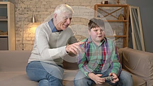 Grandfather watches as the grandson plays the console game. The old man is sitting on the couch with a young fat boy