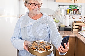 Grandma holding a lot of muffins while take her a selfie in the kitchen - woman happy and enjoyng - wearing glasses and blue