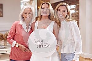 Grandmother With Adult Daughter And Granddaughter In Bridal Store Holding Yes To The Dress Sign