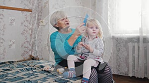 Grandmother enjoys combing hair to child on sunny morning. Good time at home. Baby sitting on granny`s laps