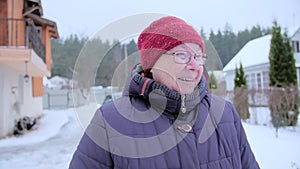 Grandmother in glasses, a red knitted hat and a blue jacket smiles at the camera.