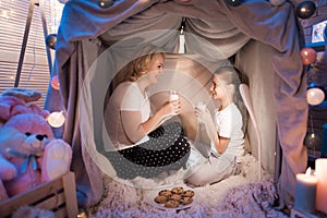 Grandmother and granddaughter are eating cookies with milk in blanket house at night at home.