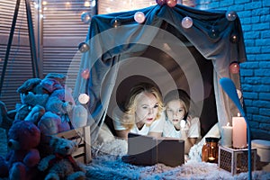 Grandmother and granddaughter are reading book in blanket house at night at home.