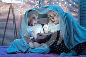 Grandmother and granddaughter are reading book with flashlight under blanket at night at home.