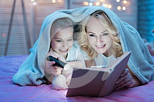 Grandmother and granddaughter are reading book with flashlight under blanket at night at home.