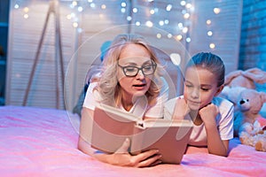 Grandmother and granddaughter are reading book before sleep at night at home.