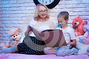 Grandmother and granddaughter are reading fairy tales book at night at home.