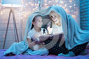 Grandmother and granddaughter are reading fairy tales under blanket at night at home.