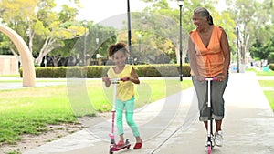 Grandmother And Granddaughter Riding Scooters In Park