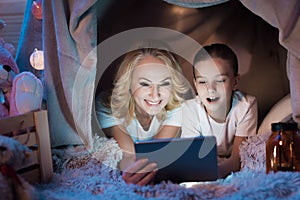 Grandmother and granddaughter are watching video on tablet in blanket house at night at home.