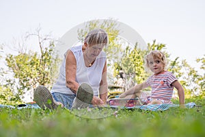 Grandmother with her grandaughter casually sitting on a blanket reading and talking in a park.