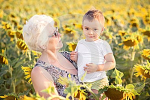 Grandmother holding in arms toddler grandson standing in the field of sunflowers in summer evening. Rustic life