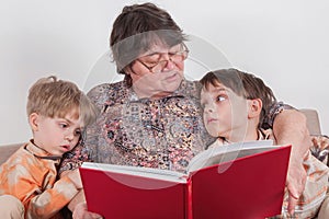 A grandmother hugs her grandchildren, who are snuggling up to her. A woman reads a book to them