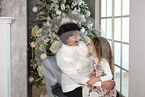 Grandmother with a little girl on the background of Christmas decorations and a large window. Family holiday, emotions, gift box.