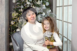 Grandmother with a little girl on the background of Christmas decorations and a large window. Family holiday, emotions, gift box.