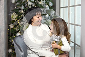 Grandmother with a little girl on the background of Christmas decorations and a large window. Family holiday, emotions, gift box.