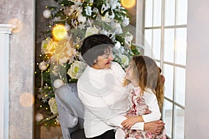 Grandmother with a little girl on the background of Christmas decorations and a large window. Family holiday, emotions, gift box.