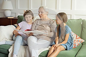 Grandmother and little granddaughters sit on sofa at home reading  book