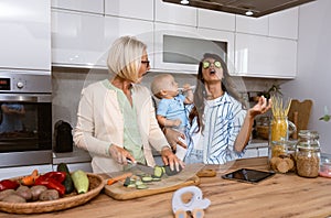 Grandmother mother and baby granddaughter cooking together in kitchen in apartment while mom making a funny faces for child and