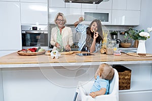 Grandmother mother and baby granddaughter cooking together in kitchen in apartment while mom making a funny faces for child and