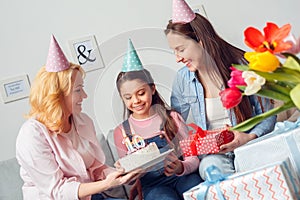 Grandmother mother and daughter together at home birthday sitting women giving girl cake and gift smiling happy