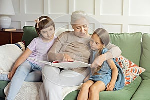 Grandmother reading aloud book to her cute little granddaughters at cozy home interior