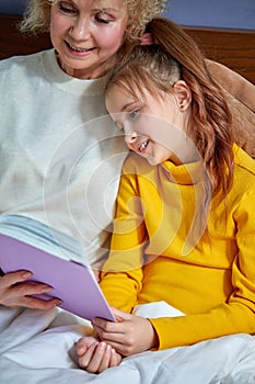 grandmother reads a book to little daughter lying on bed in children& x27;s room