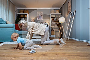 Grandmother of single mother taking care of her young baby, playing on the apartment floor. Senior woman babysitting her grandson