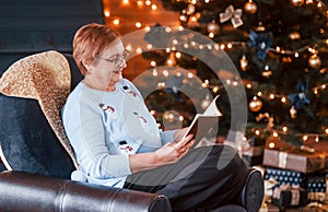 Grandmother sitting on the chair with book in festive New year room with christmas decorations