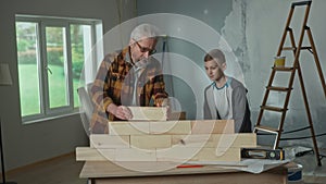 Grandpa and grandson are building a wall of wooden blocks on the table. An elderly man teaches a teenager the basics of
