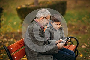 Grandpa and grandson seat on the bench and use a tablet