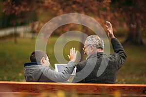 Grandpa and grandson seat on the bench and use a tablet. They put up their hand up