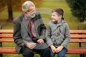 Grandpa and his grandson spend time together in the park. They are sitting on the bench and look to each other