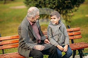 Grandpa and his grandson spend time together in the park. They are sitting on the bench and look to each other