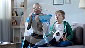 Grandpa holding Argentina flag, watching football with boy, worrying about game