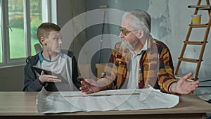 Grandpa plans renovations using a paper apartment project. The grandson helps him with the information on the tablet. An