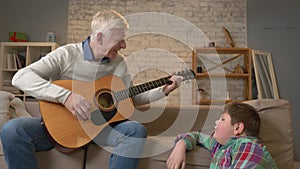 Grandpa plays for his grandson on guitar, music. A young fat guy is sitting on the haven with his grandfather. Teaches