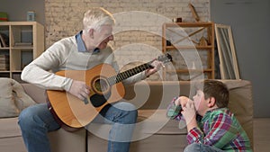 Grandpa plays for his grandson on guitar, music. A young fat guy is sitting on the haven with his grandfather. Teaches