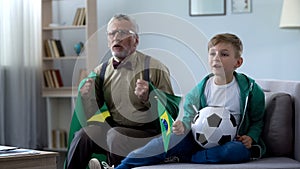 Grandpa wrapped in Brazil flag watching football with boy, worrying about game