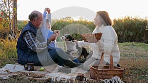 Grandparent with grandchild are sitting on blanket near pet on picnic against autumn nature background