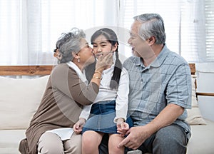 Happy asian grandparents sitting on sofa at home playing and kiss on cheek of little granddaughter