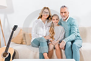 grandparents embracing smiling granddaughter and looking at camera on sofa