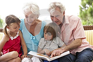 Grandparents And Grandchildren Reading Book On Garden Seat