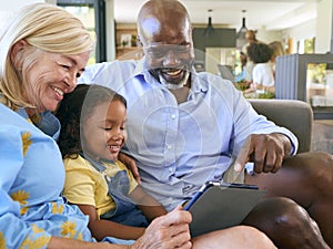 Grandparents And Granddaughter With Digital Tablet Sitting On Sofa At Home Together