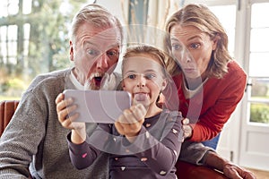 Grandparents With Granddaughter Pulling Faces Posing For Selfie On Mobile Phone At Home Together