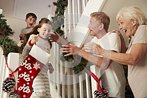 Grandparents Greeting Excited Grandchildren Wearing Pajamas Running Down Stairs Holding Stockings On Christmas Morning