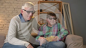 Grandson feeds his grandfather with chips from his hand. Grandfather and grandson are sitting on the couch and watching